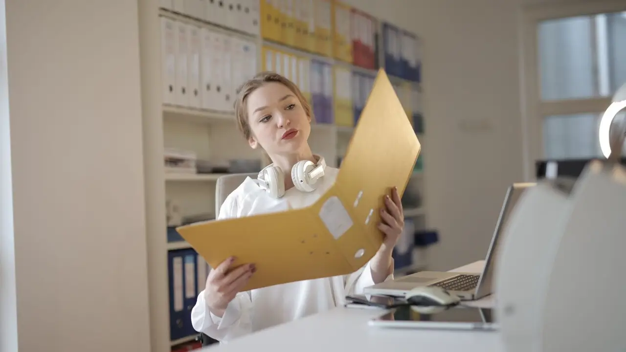 Joven trabajadora consultando una carpeta en una oficina con estanterías de archivo.